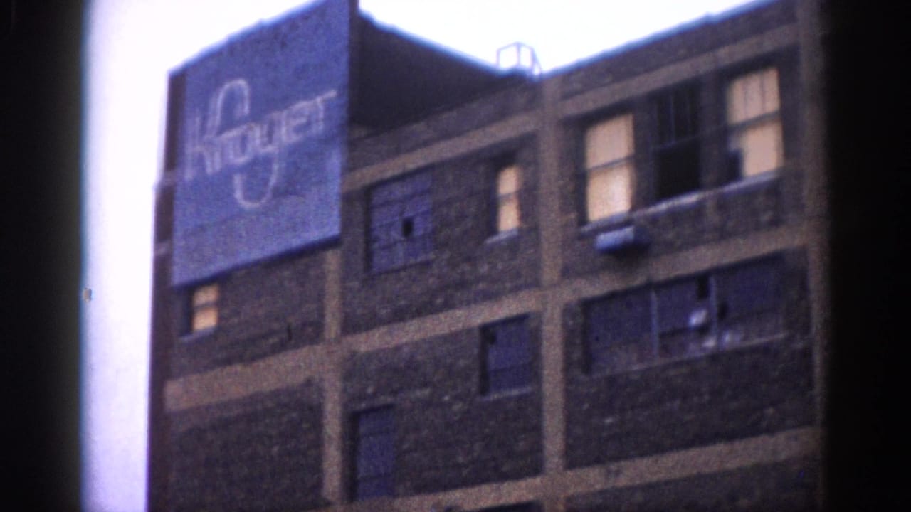 Multi-story brick commercial building with a Kroger sign on its upper facade, viewed from street level at the start of a demolition sequence.