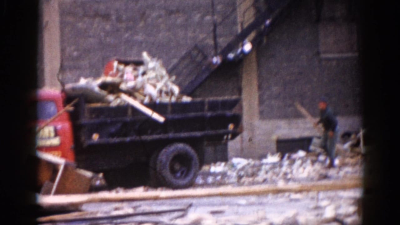 Red dump truck with its bed loaded with wooden beams and demolition rubble, parked beside a partially demolished brick building; one worker visible at right.