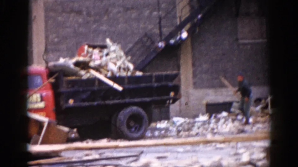 Red dump truck being loaded with lumber and brick rubble at the Illinois demolition site circa 1962; a worker in period work clothes stands on the debris pile at right.
