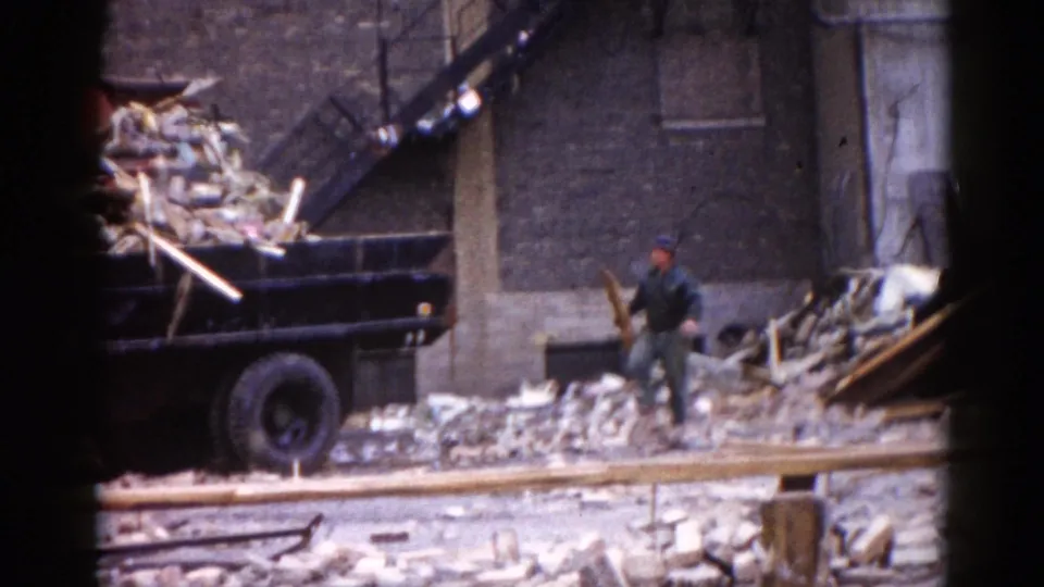 Demolition worker in period work clothes clearing rubble at the Illinois site circa 1962; brick and lumber debris fills the foreground with what appear to be construction rail tracks.