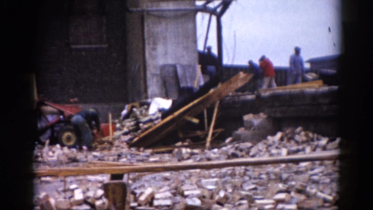 Workers and a small construction vehicle amid brick rubble and angled beams at a clearance site