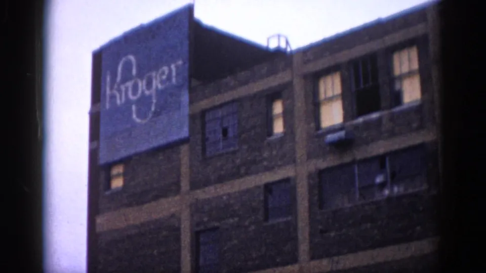 Upward view of a multi-story brick warehouse or commercial building bearing a large painted Kroger sign, consistent with early 1960s Illinois urban context, with some window panes appearing damaged or missing.