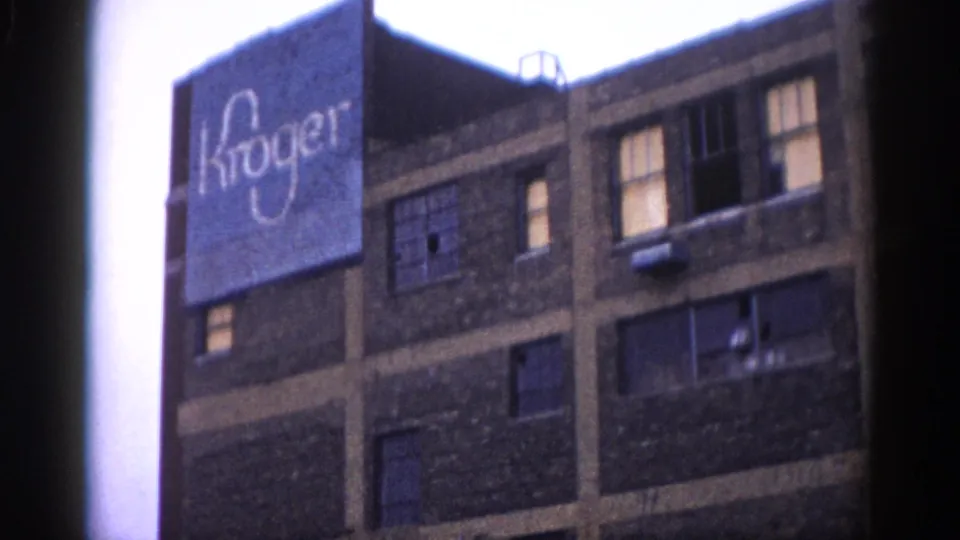 Close upward view of a multi-story brick building displaying a large painted Kroger sign, likely a warehouse or distribution facility in Illinois circa 1962, with rows of windows and an overcast sky above.