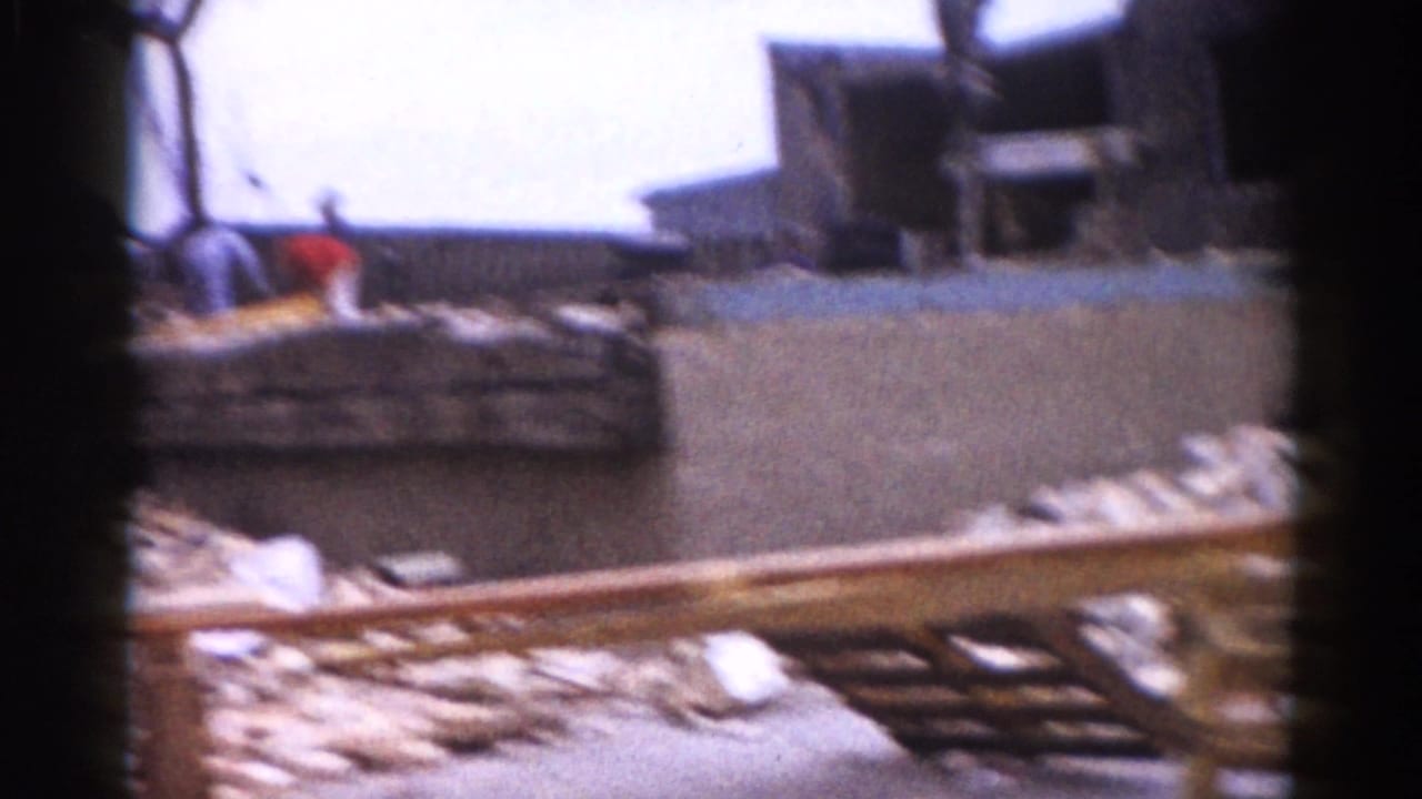 Two workers near a close pile of broken masonry and lumber; one wears a red shirt, the other lighter clothing.