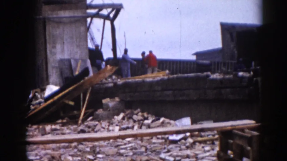 8mm color footage, c.1962 Illinois — two workers, including a man in a red shirt, stand among rubble; concrete pillars and an industrial building remnant rise behind them.