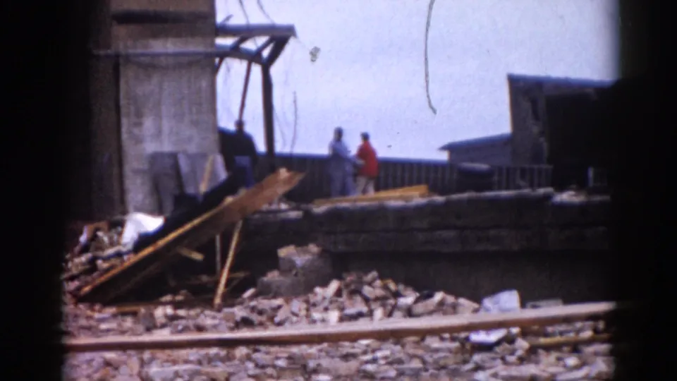 8mm color footage, c.1962 Illinois — man in red shirt and a companion stand amid rubble; concrete piers and scattered masonry fill the midground under an overcast sky.