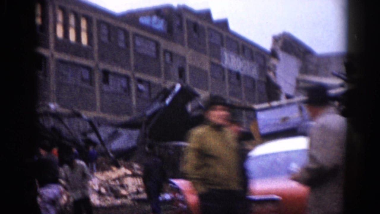 Close-up of debris-strewn street with people in period clothing near a partially visible red vehicle, damaged multi-story brick building behind.