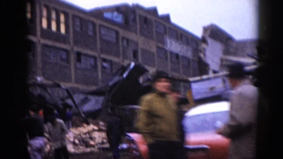 8mm home movie frame, c.1962, Illinois: two bystanders in period coats move through rubble beside a partial red vehicle, large damaged brick building visible behind.