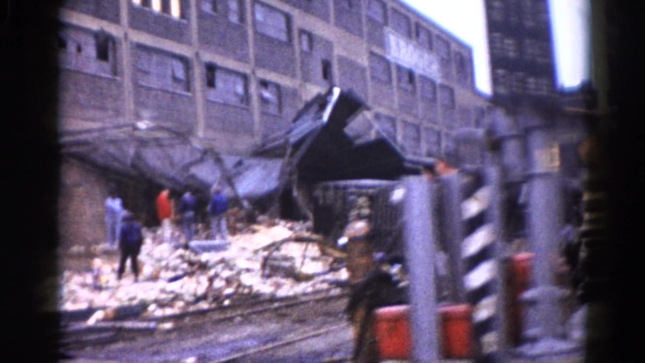 8mm home movie, c.1962, Illinois: collapsed multi-story brick building with facade destroyed, rubble spread across street, bystanders including a person in red visible near a utility pole.