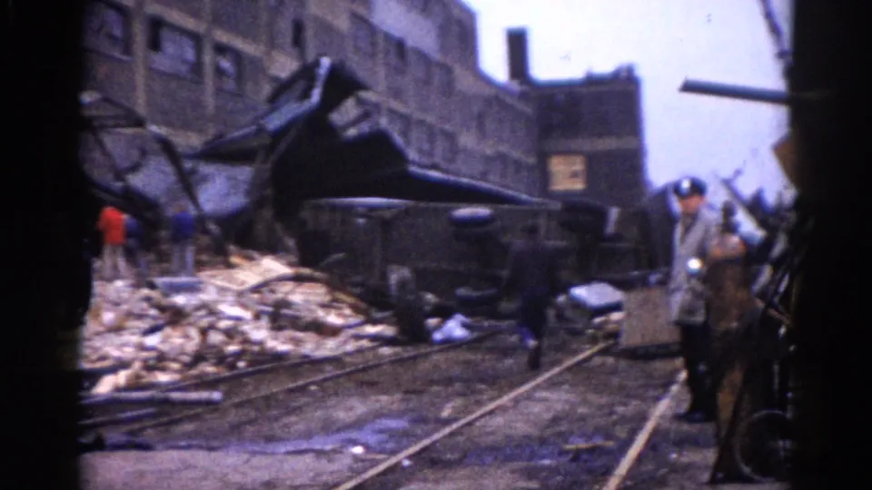 8mm home movie, c.1962, Illinois: railroad or streetcar tracks exposed in a rubble-covered street, twisted metal wreckage, an apparent uniformed official moves among bystanders.
