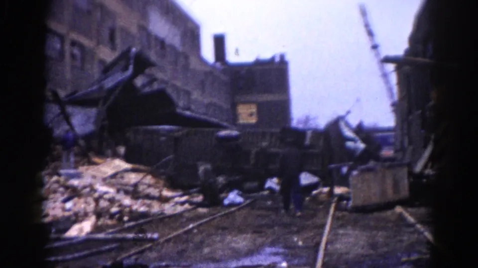 8mm home movie, c.1962, Illinois: broad aftermath view showing twisted metal wreckage, exposed railroad tracks, a cylindrical drum or barrel, and damaged background buildings under overcast sky.