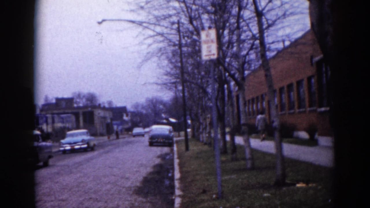 8mm home movie, Illinois c. 1962: tree-lined suburban street with parked early-1960s American cars and a multi-story brick building at right, small figures visible in the background.