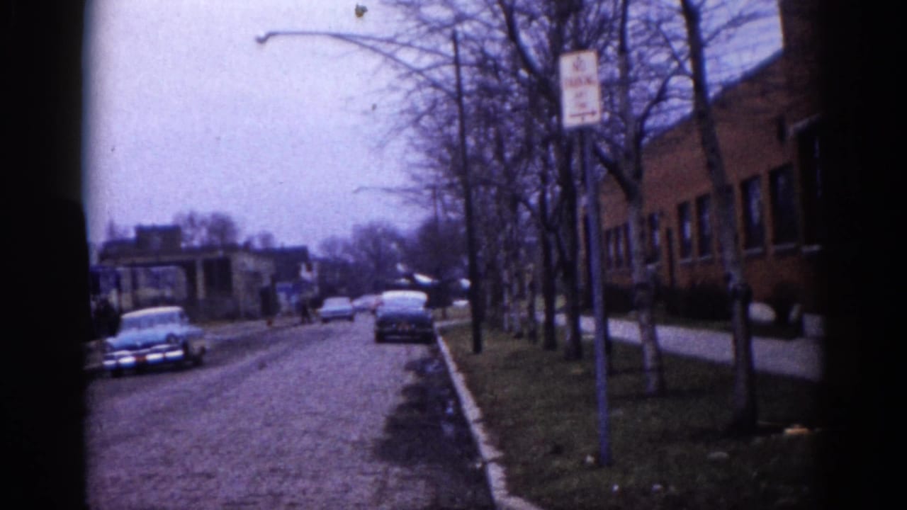 Similar suburban street angle with a dark American sedan at the curb, bare trees, brick building at right, and a street lamp pole in the foreground.