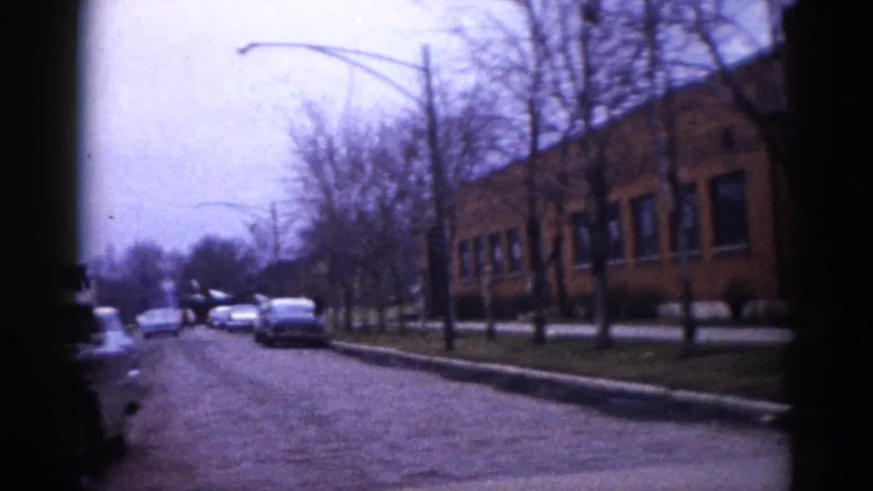 8mm home movie, Illinois c. 1962: panning shot of a large brick building facade along a bare-treed suburban road with parked early-1960s cars and overcast winter sky.