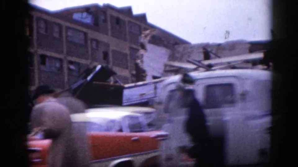 8mm home movie, c. 1962: abrupt handheld pan reveals apparent structural damage on a brick building, with a red vehicle, white work truck, and two unidentified standing figures.