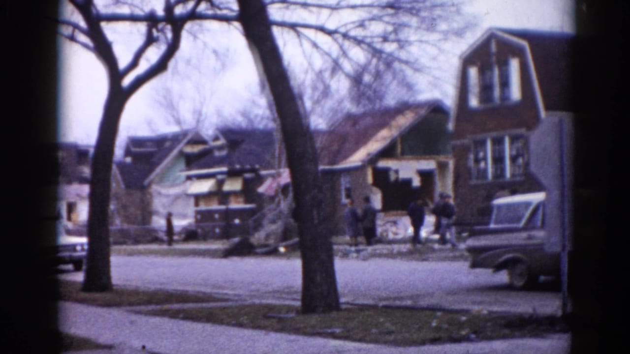 Residential street with bare winter trees, early 1960s vehicles, and people near a house under apparent construction or renovation.