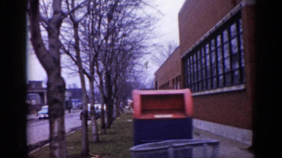 Mid-century modern brick school building with curtain-wall windows in an Illinois neighborhood, circa 1962, with a red postal collection box and parked cars visible on the street.