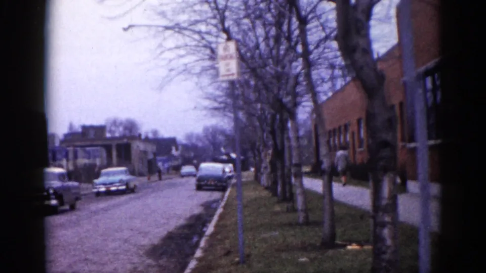 Illinois neighborhood street circa 1962 looking along the block past the brick school building, with early 1960s cars, bare trees, a street sign, and a large vehicle visible in the distance.