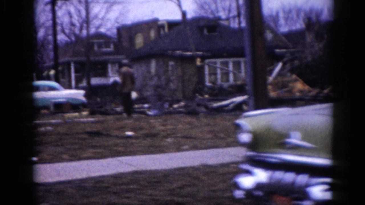 Circa-1962 Illinois suburban lot showing a collapsed or heavily damaged residence, debris scattered across the yard, and a late-1950s American sedan at frame left.