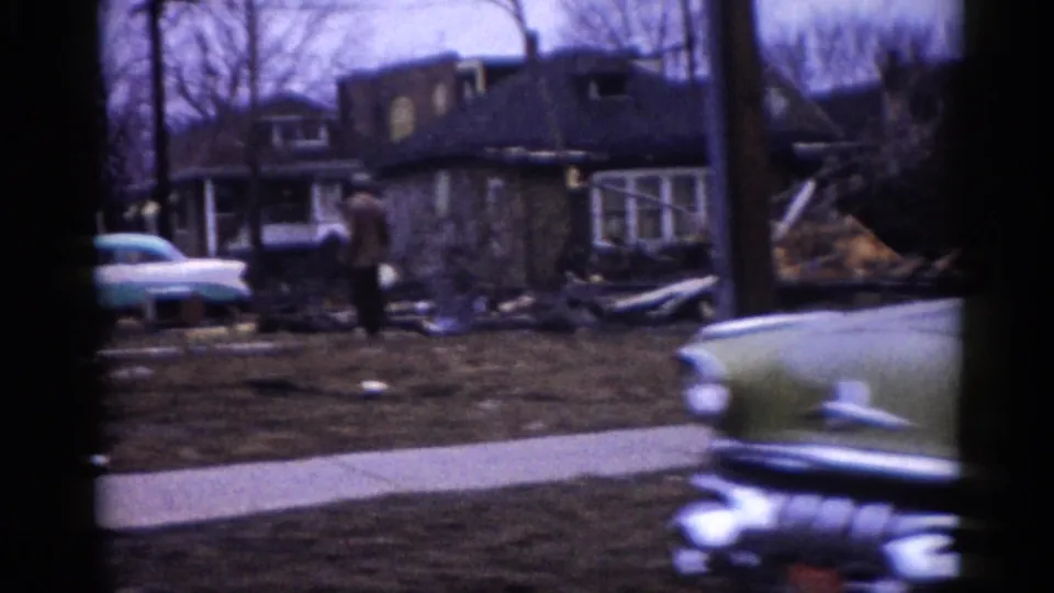 Circa-1962 Illinois suburban lot showing a collapsed or heavily damaged residence, debris scattered across the yard, and a late-1950s American sedan at frame left.
