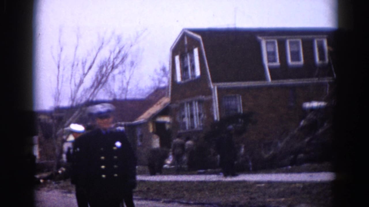 Uniformed police officer standing in foreground; brick two-story house at right; debris and bystanders visible behind.