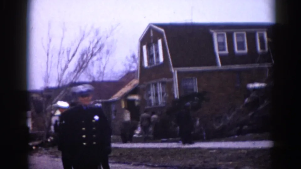 A uniformed police officer with cap and brass buttons stands watch near a damaged structure in what appears to be a circa-1962 Illinois suburban neighborhood.