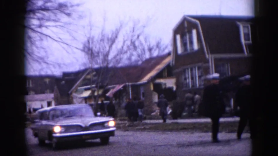 A circa-1962 Illinois residential street showing a mix of intact and damaged two-story homes, an early-1960s sedan with headlights on, and figures gathered on the roadway.