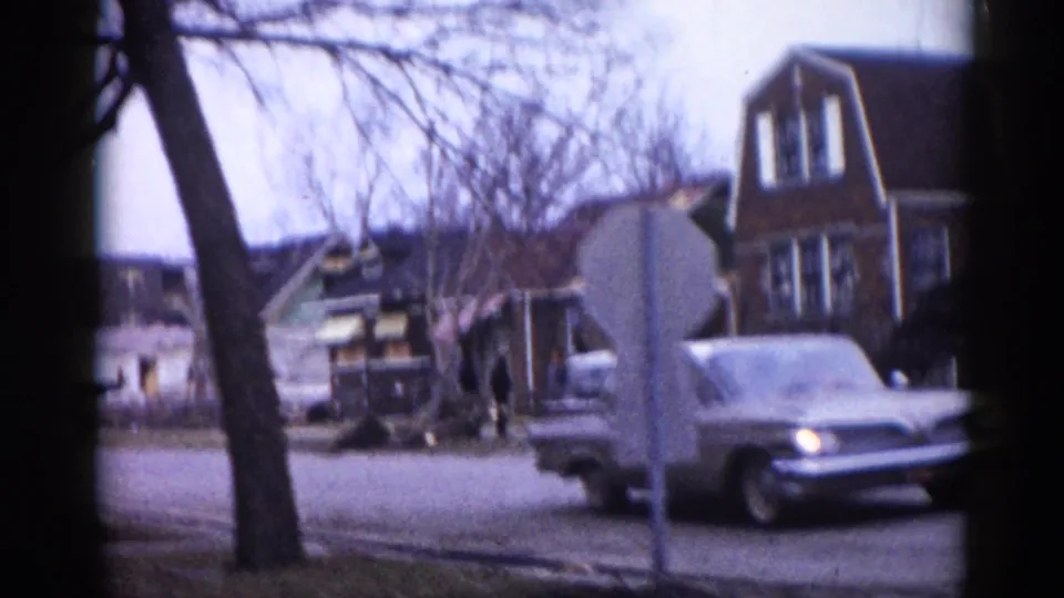 A circa-1962 Illinois suburban street shows a white van or light truck at right, bare trees, two-story brick homes, and damaged structures visible at left edge.