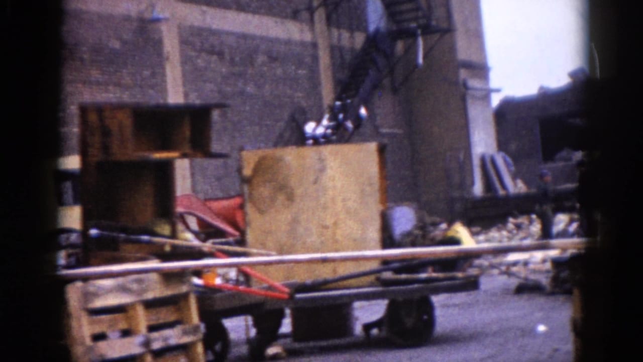 Close-up of wooden pallets, stacked metal pipes, and a red cart amid scattered construction debris, with a brick wall behind.