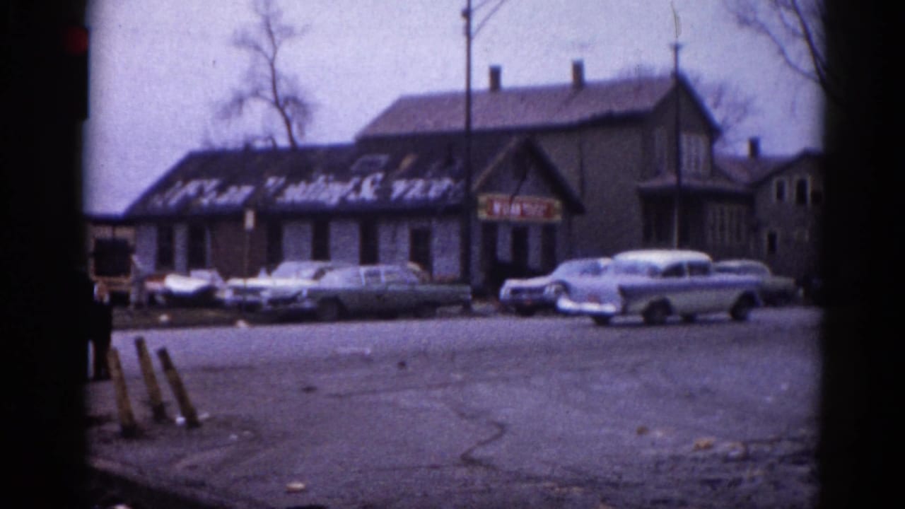 Small-town commercial building with faded painted window lettering and a row of classic American sedans parked in front, 8mm home movie footage, Illinois circa 1962.