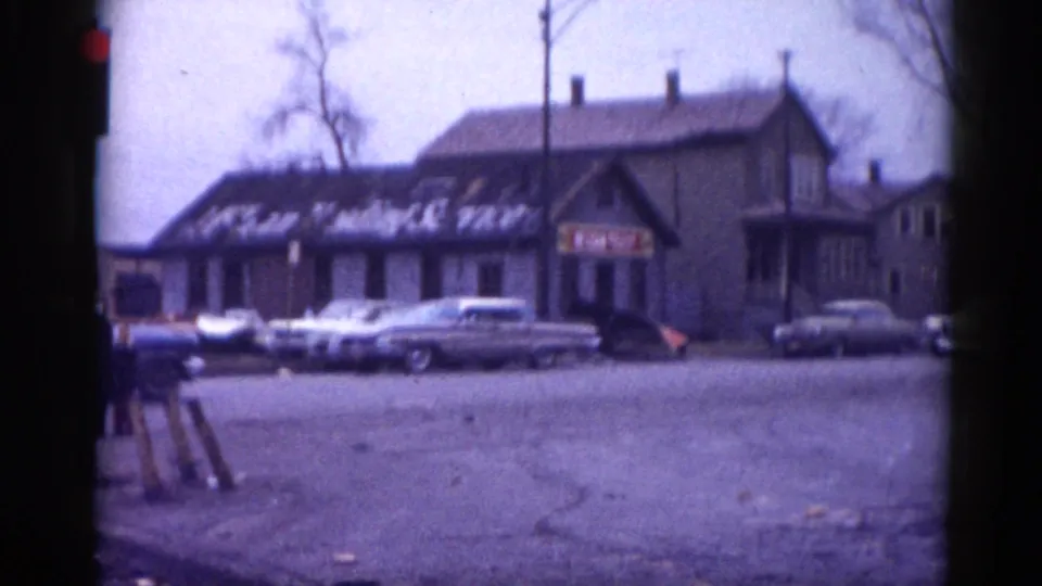 Multiple early-1960s American sedans parked across an unpaved lot from a small commercial building with hand-painted window signage, 8mm footage, Illinois circa 1962.