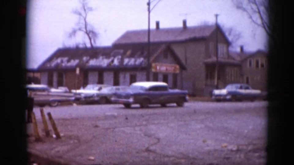 Early-1960s American cars parked outside a small-town commercial building with painted signage; wooden sawhorses appear in the right foreground, 8mm footage, Illinois circa 1962.