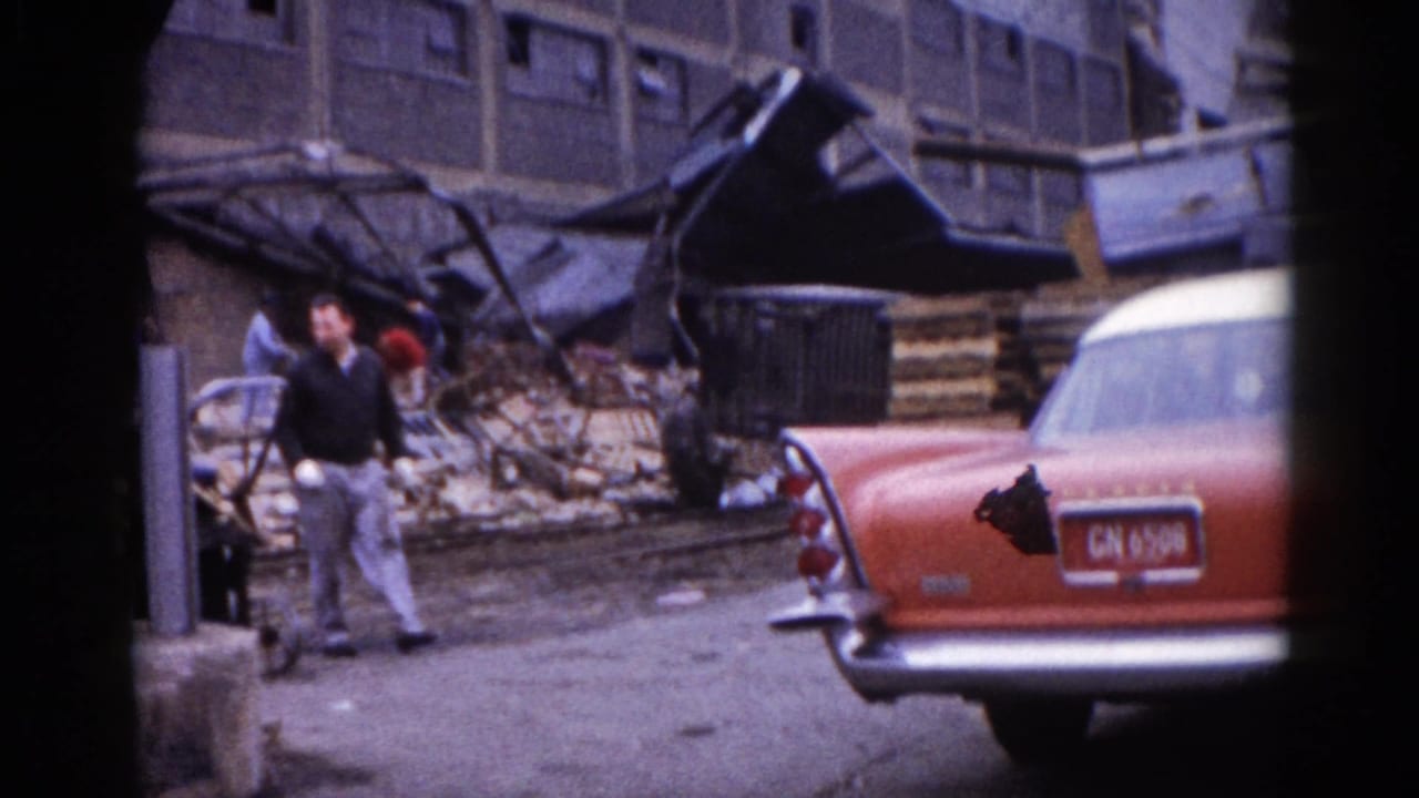 Man in dark sweater walks past a red-and-white American sedan on a rubble-strewn street; twisted metal and a collapsed structure visible behind him.
