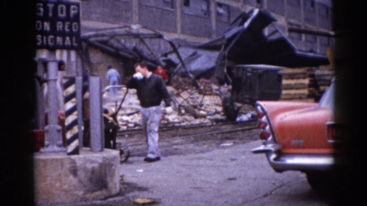 Man in dark sweater stands near striped bollards amid debris; 'STOP ON RED SIGNAL' sign visible upper right; red classic sedan and collapsed structure behind.
