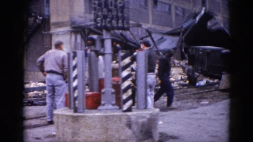 Multiple men navigating post-disaster rubble on an Illinois street circa 1962, with striped bollards, a red bucket, and a large brick building in background.