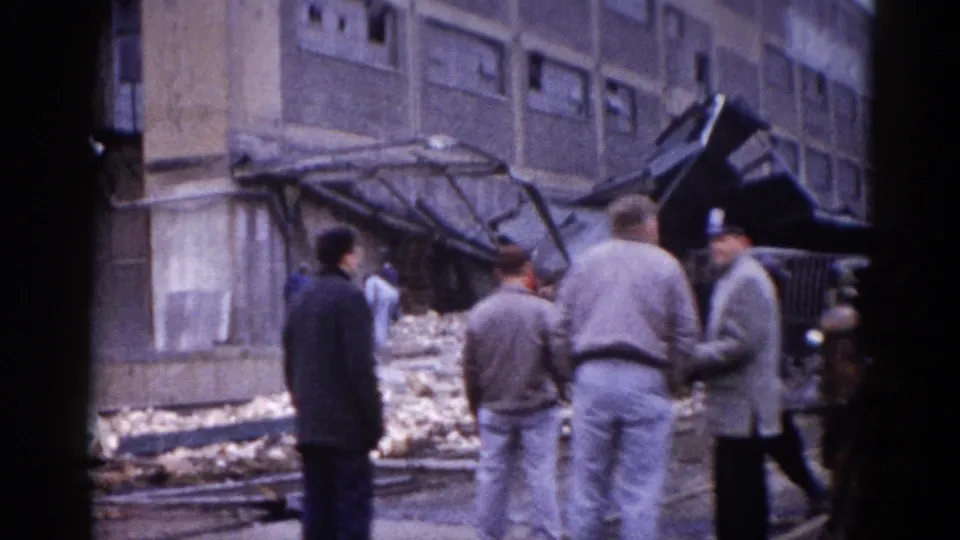 Three adult men conversing as observers of disaster aftermath, Illinois circa 1962; large brick building and collapsed structure behind them.
