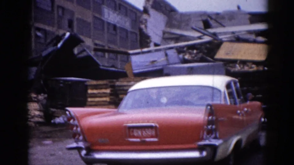 Two-tone red-and-white American sedan in foreground with legible license plate; a person stands among the debris of a collapsed building, Illinois, circa 1962.