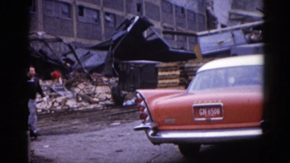 Close view of the tail-fin rear of a two-tone American sedan with a legible license plate, parked beside building collapse debris, Illinois, circa 1962.