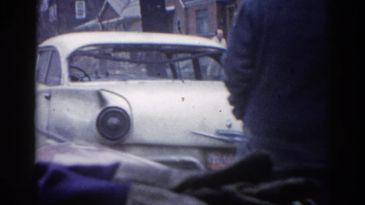 White classic American sedan on a residential street, silhouetted dark-coated figure in foreground, brick buildings behind.
