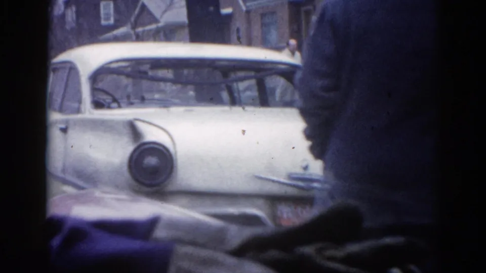 8mm home movie frame, Illinois 1962: white early-1950s-style American sedan on a residential street, dark-coated figure partially blocking view, brick buildings in background.