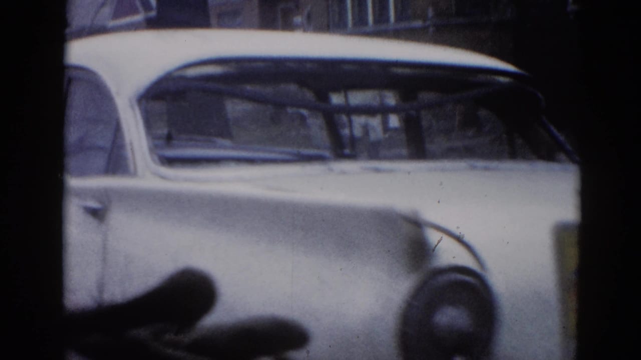 8mm film frame circa 1962 Illinois: passenger-side view of white classic American sedan with rounded roofline and chrome trim, brick residential buildings in background.