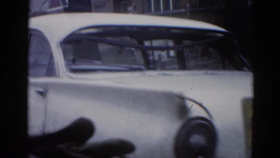 8mm film frame circa 1962 Illinois: passenger-side view of white classic American sedan with rounded roofline and chrome trim, brick residential buildings in background.