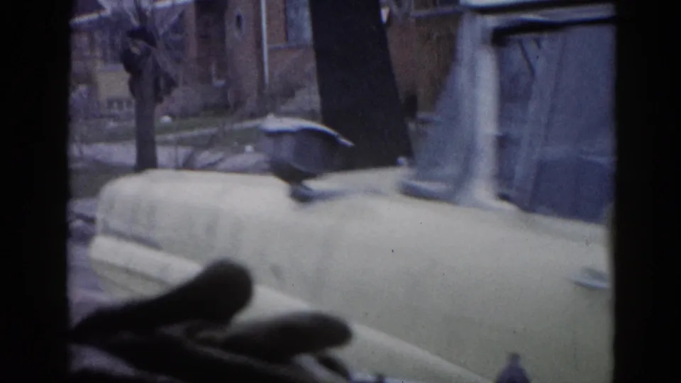 8mm home movie frame circa 1962 Illinois: camera panning past a vehicle roofline toward a brick residential building and bare winter trees.