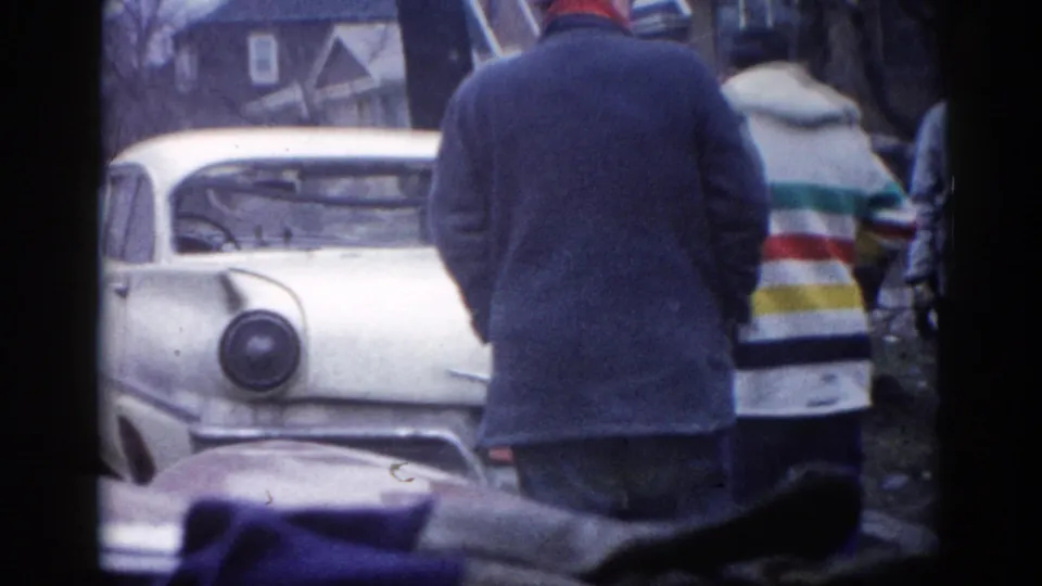 Man facing away stands beside a white early-1960s American sedan during storm cleanup, person with striped jacket partially visible at right, residential structures behind