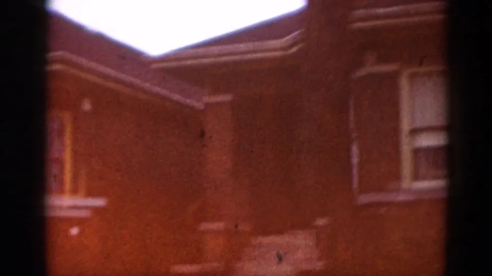 8mm film frame circa 1962 showing the roofline and upper facade of a brick house in Illinois, shot at a low upward angle with warm reddish tones.