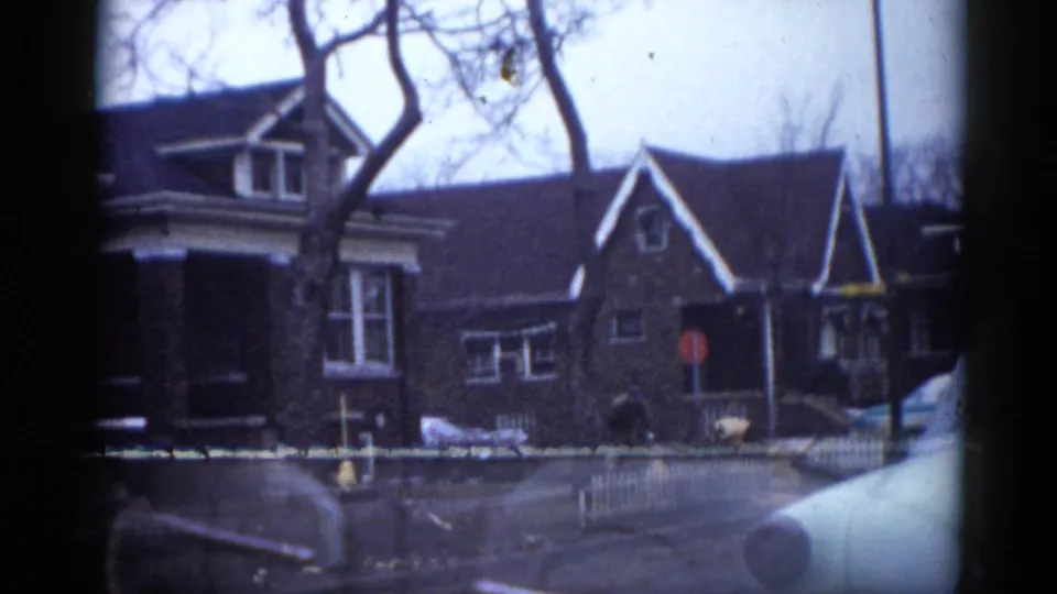 Street-level view of a brick residential building with bare trees, white picket fence, and parked car in an Illinois suburban neighborhood, 1962 8mm film.