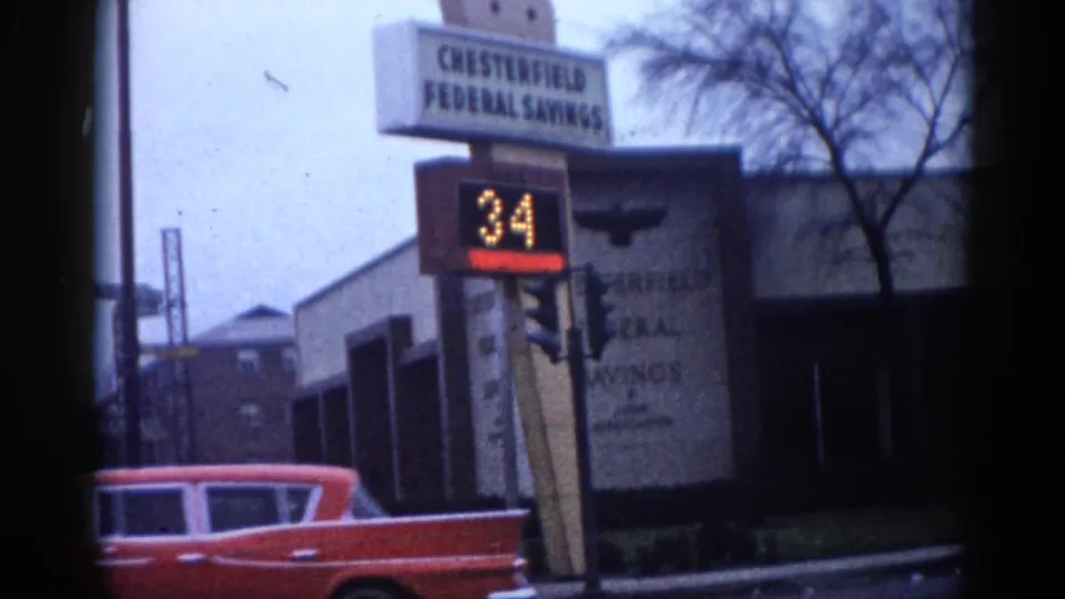 8mm home movie frame from what appears to be early-1960s Illinois: Chesterfield Federal Savings bank with LED sign showing temperature 34 and a red classic American car in the left foreground.