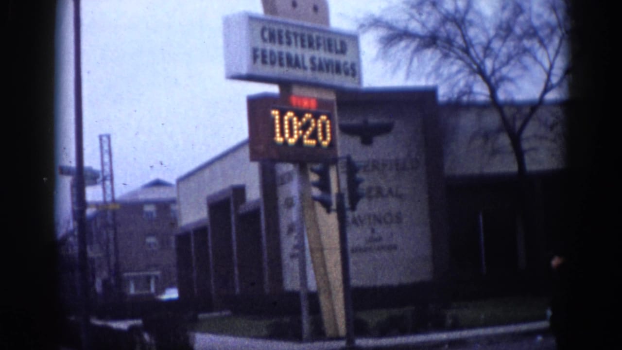 8mm home movie frame from what appears to be early-1960s Illinois: Chesterfield Federal Savings bank with LED sign cycling to time 10:20 and a traffic light at the street corner.
