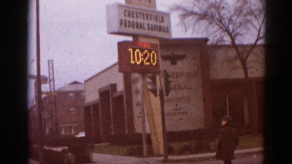 8mm home movie frame from what appears to be early-1960s Illinois: Chesterfield Federal Savings bank with LED display at 10:20 and a pedestrian in dark clothing crossing the foreground sidewalk.