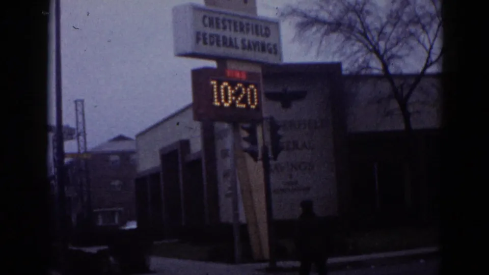 8mm home movie frame from what appears to be early-1960s Illinois showing Chesterfield Federal Savings bank, LED sign at 10:20, and bare trees under a blue-tinted overcast sky.